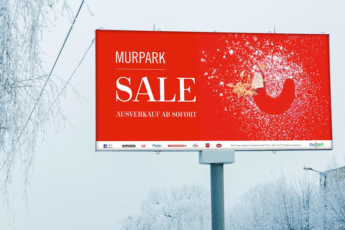 Zu sehen ist ein großes Plakat der Murpark Weihnachts-Kampagne in verschneiter Landschaft. Auf dem Plakat ist ein fast komplett aufgegessenes Vanillekipferl abgebildet und es steht "Sale - Ausverkauf ab sofort!" geschrieben.