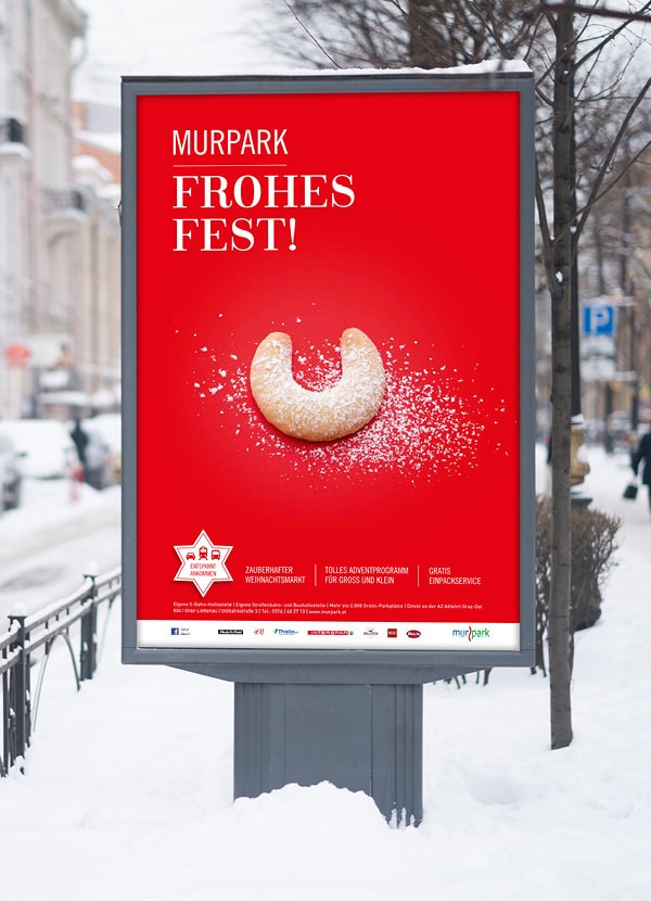 Zu sehen ist ein Plakat der Murpark Weihnachts-Kampagne in verschneiter Innenstadt-Landschaft. Auf dem Plakat ist ein Vanillekipferl abgebildet und es steht "Frohes Fest!" geschrieben.