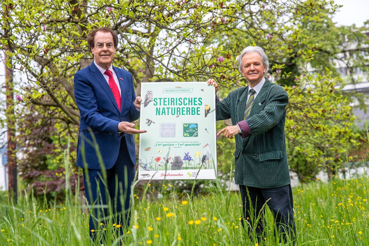 Auf dem Foto sind Prof. Dr. Johannes Gepp und Hans Roth zu sehen. Sie halten ein Plakat der Kampagne "1m2 genützt - 1m2 geschützt" des steirischen Naturerbes.